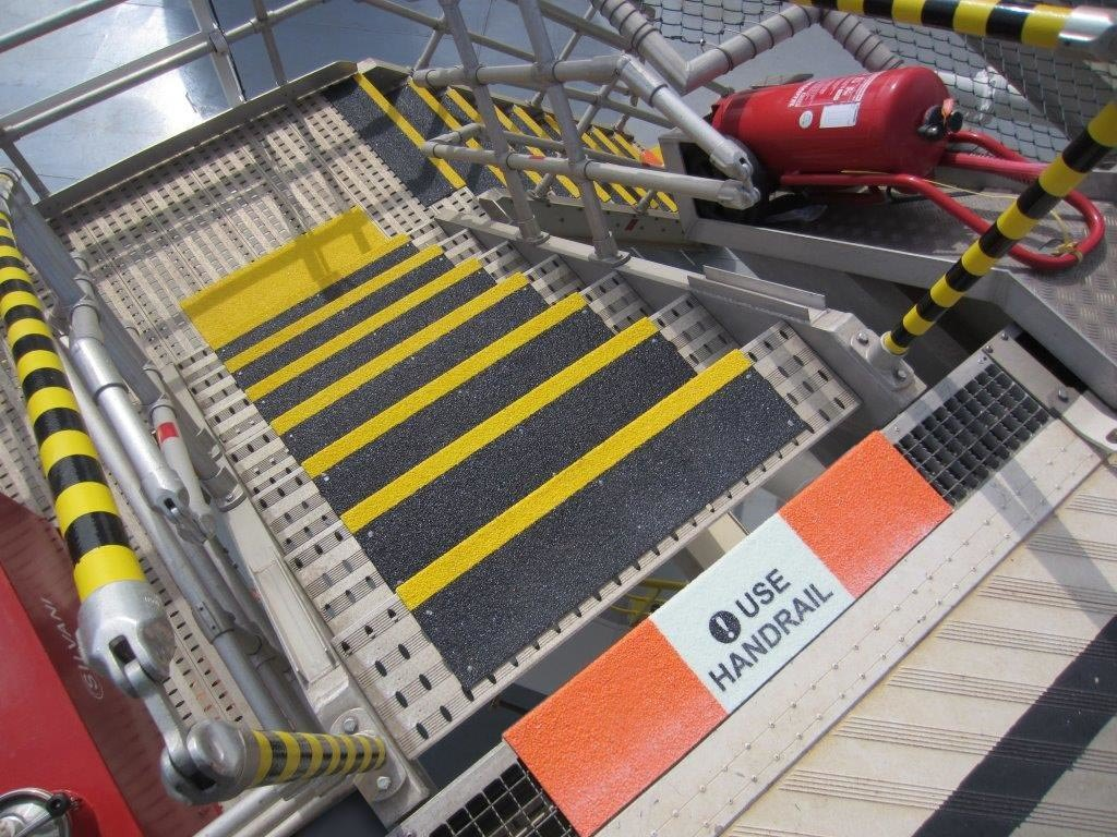 Two-tone yellow/black step covers on fabricated metal stairs, a walkway cover at the bottom, and a glow-in-the-dark “Use Handrail” message at the top